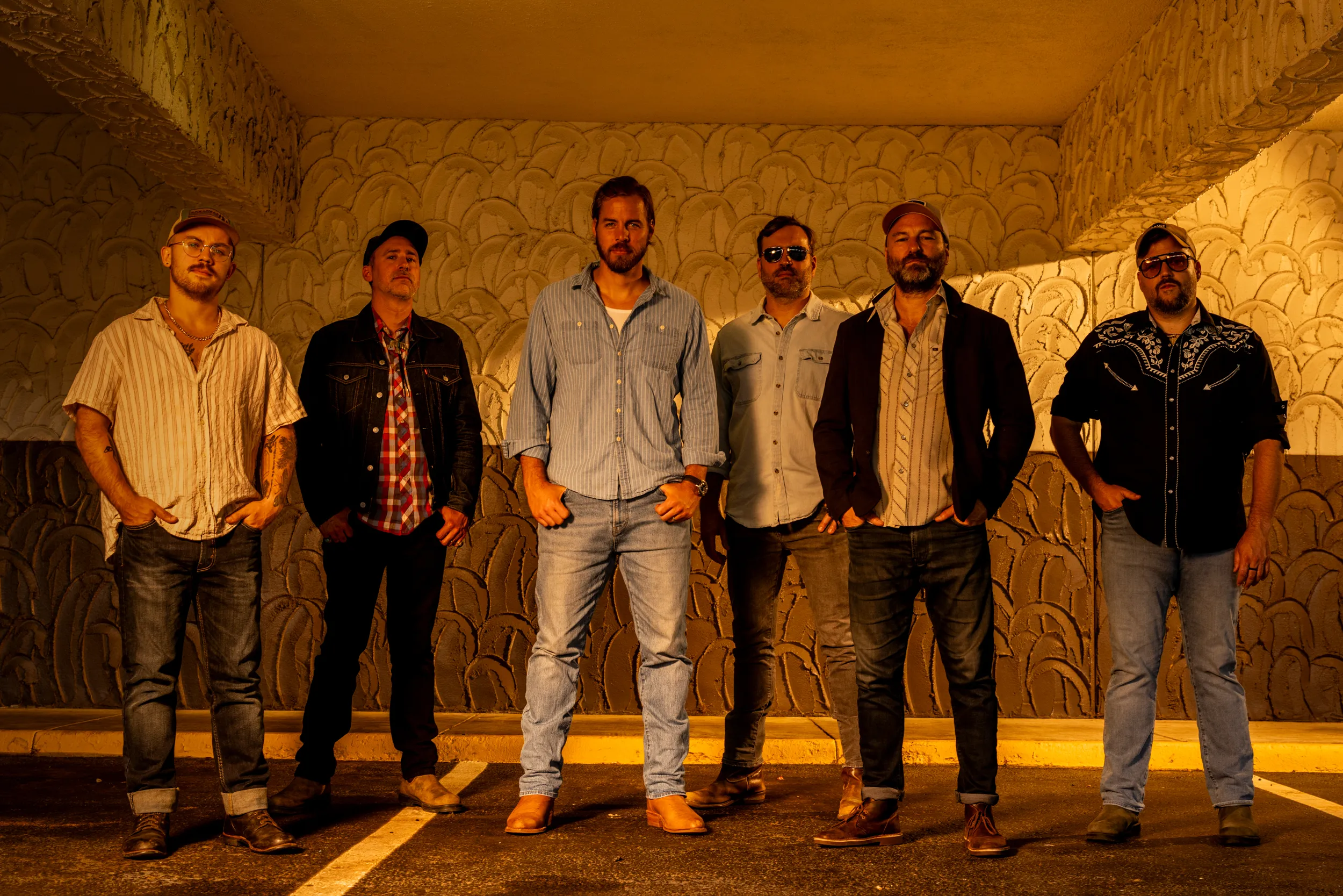 The six members of Big Holler standing shoulder to shoulder against a scalloped concrete wall under warm sodium light